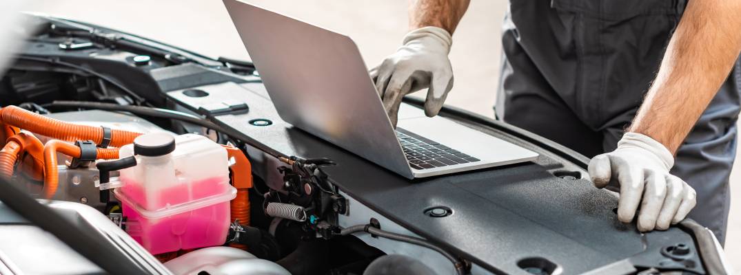 Mechanic standing in front of a car with the hood open, looking down at a laptop.