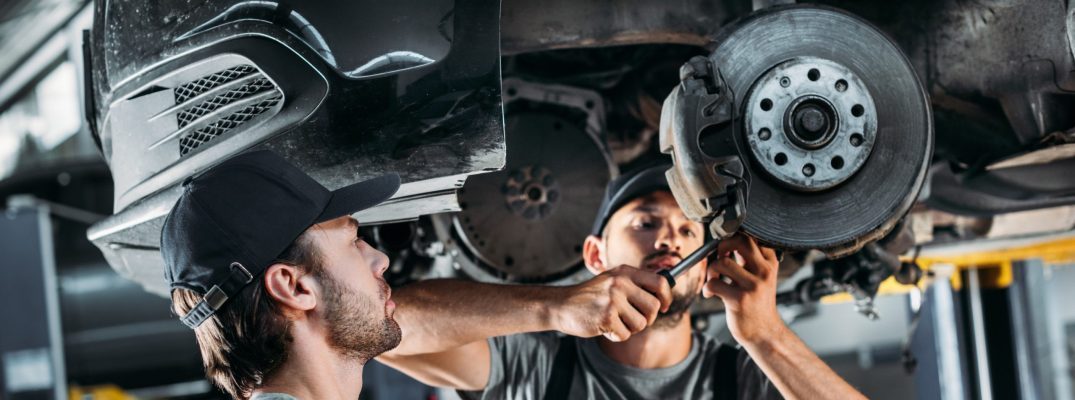 Two mechanics look at a car's brake pads from below.