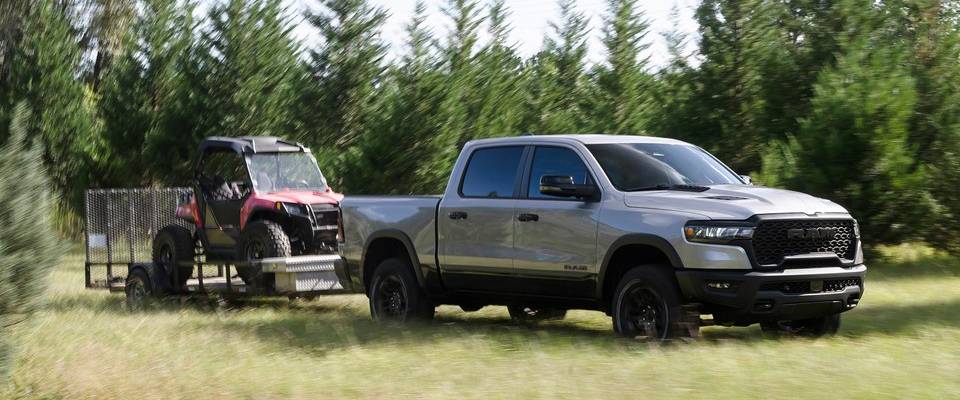 A silver 2026 Ram 1500 pulls a trailer with a red buggy on it across a green field with trees in the background