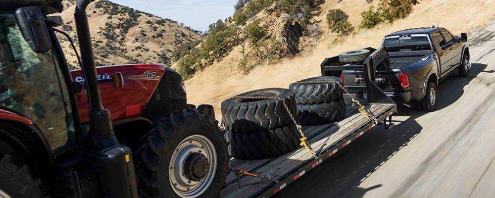 A black 2026 Ram HD pulls a trailer loaded with large tires and a red tractor. New 2026 Ram 2500 & 3500 Towing and Payload Guide at Blenheim Chrysler Dodge Jeep Ram in Blenheim, ON