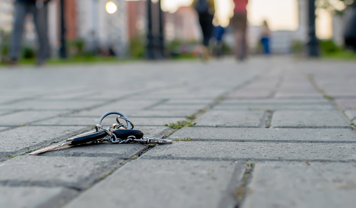 A set of car keys lies on a paved walkway, slightly tilted with a key fob and multiple keys attached to a metal ring. In the blurred background, people are walking along the path, unaware of the lost keys.