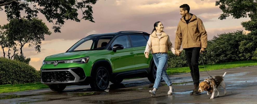 A happy couple and their dog pass a green 2025 Volkswagen Taos that is parked in front of a sunset