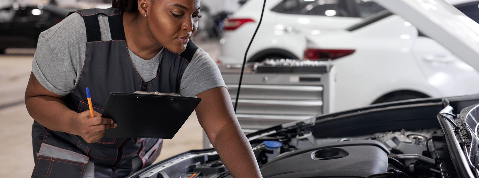 car service, job and profession concept. young african female worker in gray uniform with clipboard and pen writing over auto repair shop on background