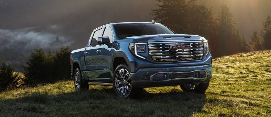 A blue 2026 GMC Sierra 1500 is parked in front of a sunset on a grassy hill