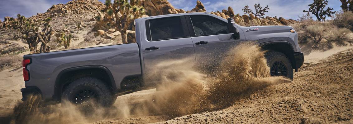 A silver 2025 Chevrolet Silverado HD drives, kicking up sand in front of a rocky backdrop