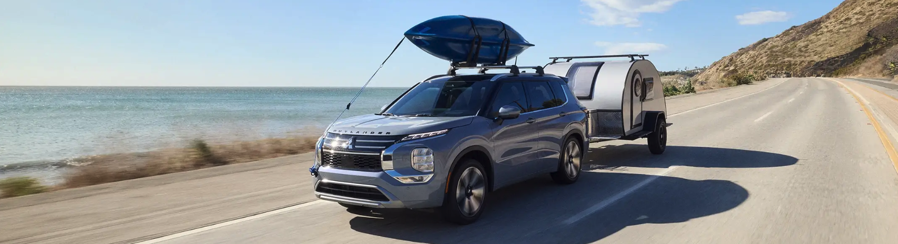 Outlander with canoe on roof racks, towing a mini camper on road with body of water in background