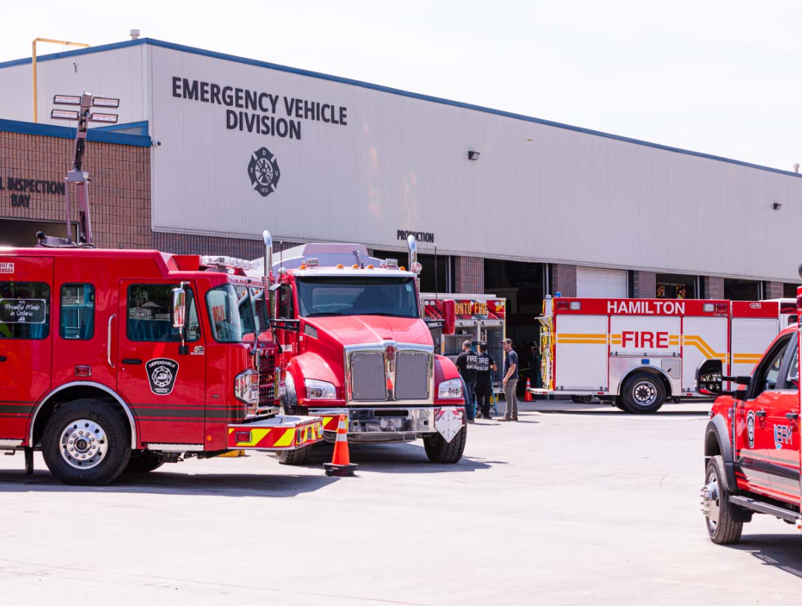 multiple_red_fire_trucks_parked_outside_emergency_vehicle_building_with_garage_doors_open_and_personnel_visible@2x