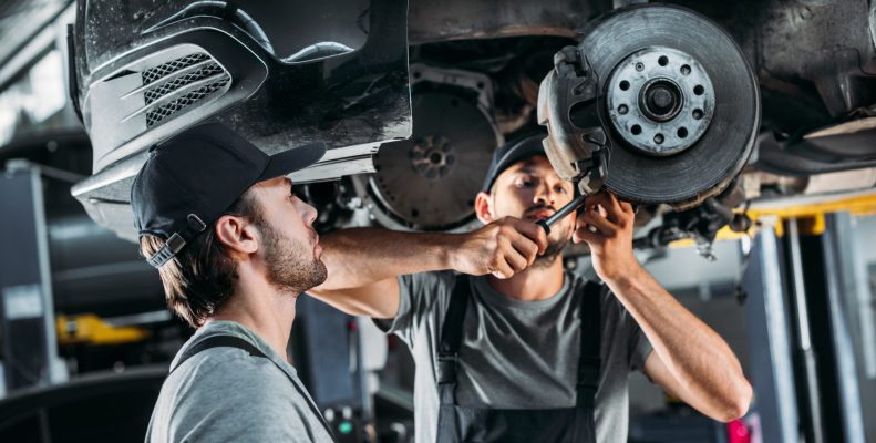 Two mechanics work on the underside of a vehicle.