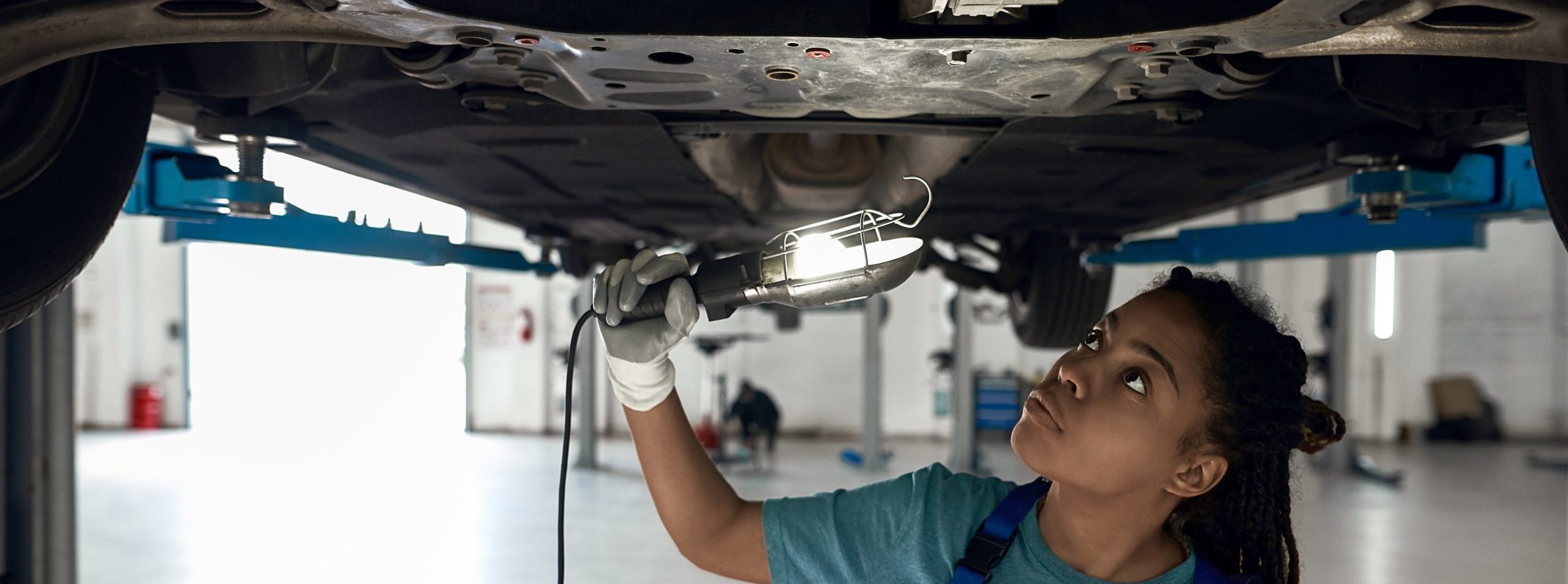 Young afro american girl checking bottom of car installed on lift at car service station with flashlight.