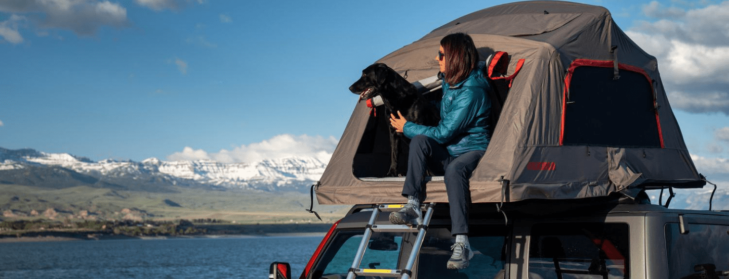 Unknown person sitting inside tent on top of Ford vehicle with her dog near a body of water, looking out into the horizon