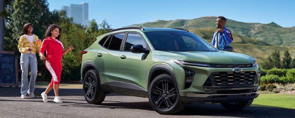 A green 2026 Chevrolet Trax is parked outside a cabin with green hills in the background. A group of people are walking towards it.
