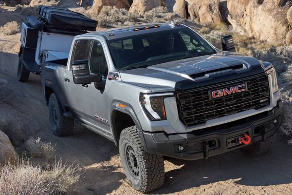A silver 2026 GMC Sierra HD pulls a trailer on a rocky off-road trail. New 2026 GMC Sierra 2500 & 3500 towing and engine specs at Gardner GM in Hope, BC