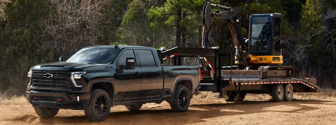 A black 2026 Chevrolet Silverado HD pulls a CAT vehicle on a trailer across a worksite. New 2026 Chevrolet Silverado 2500 & 3500 engine and towing specs at Gardner GM in Hope, BC