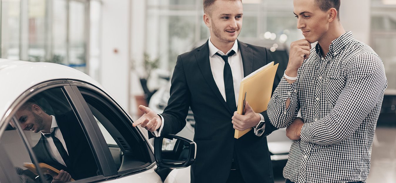 Professional car dealer showing cars on sale to his male customer, working at the auto dealership salon. Man looking thoughtfully at the car he is examining. Handsome man buying new automobile