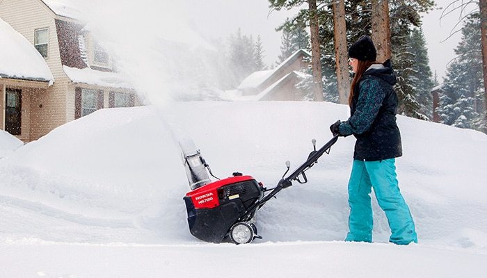 A woman using a single stage snowblower in front of a house by a mound of snow