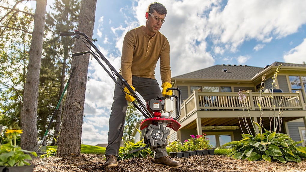 Man bending over and using a tiller in a backyard with a house in the background