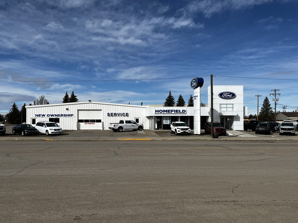 Exterior view of Homefield Ford dealership in Southern Alberta with Ford signage, service department, and multiple vehicles parked in front under a blue sky.