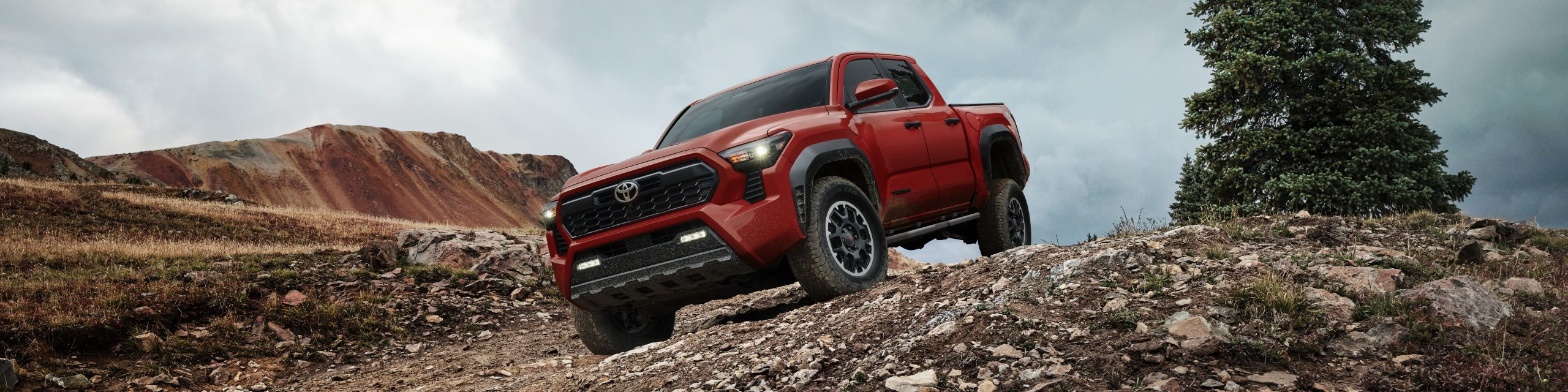 Red Toyota Tacoma TRD Off-Road Premium driving on rocky mountain terrain with cloudy sky background.