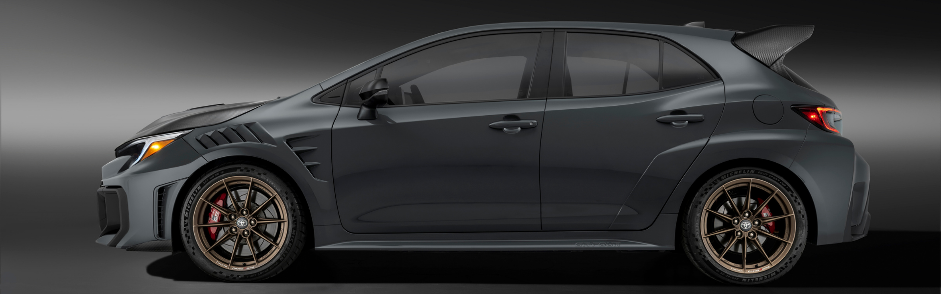 Side profile of a dark grey Toyota GRMN Corolla with bronze wheels and rear spoiler
