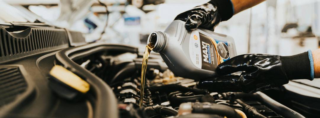 A pair of gloved hands pours oil from a silver bottle into a car's engine