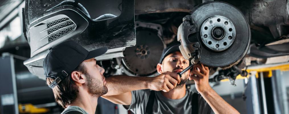 Two technicians work on the brakes of a car that has been hoisted up to let them access the underside