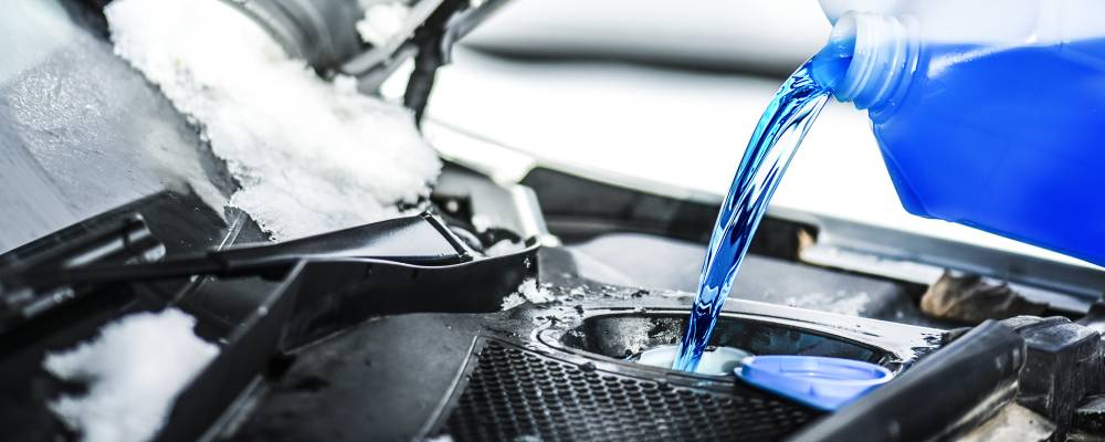 A clear bottle pours a blue antifreeze/coolant mixture into a car's reservoir