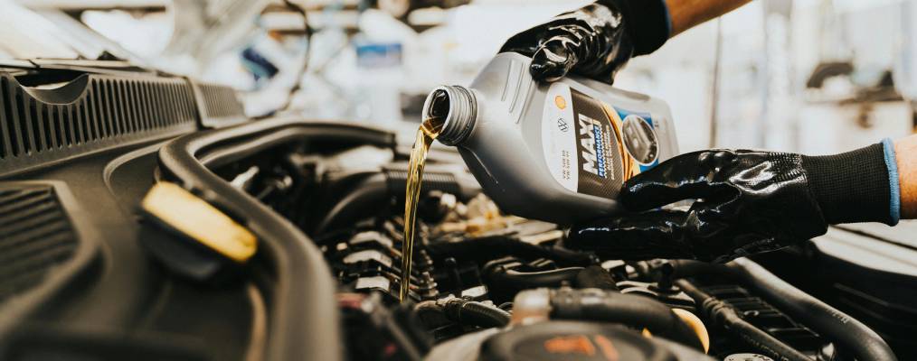 Gloved hands pour motor oil from a silver bottle into a car's reservoir