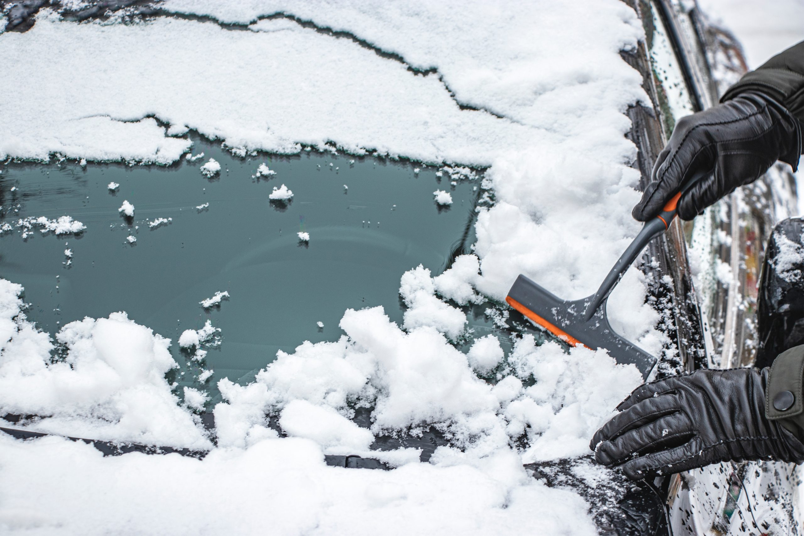A hand uses a scraper to clear ice and snow from a windshield