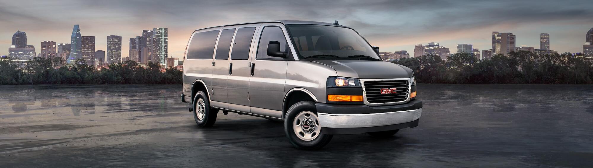 A silver 2025 GMC Savana Passenger or Cargo van sits in front of a low-light cityscape