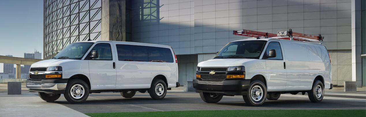 Two white 2025 Chevrolet Express Passenger & Cargo Vans in the parking lot outside of a white and glass building