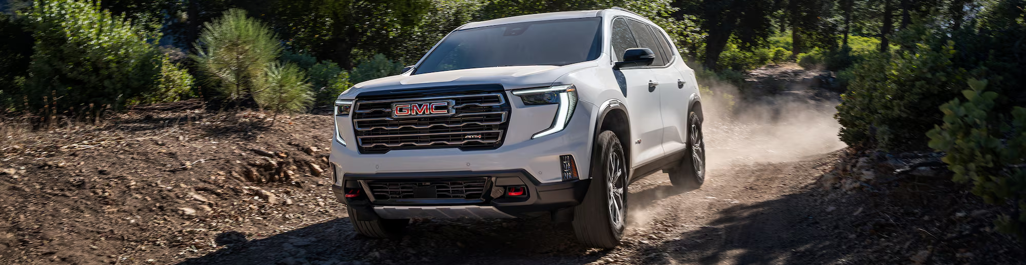 A white 2026 GMC Acadia drives over dirt road at an angle approaching the camera