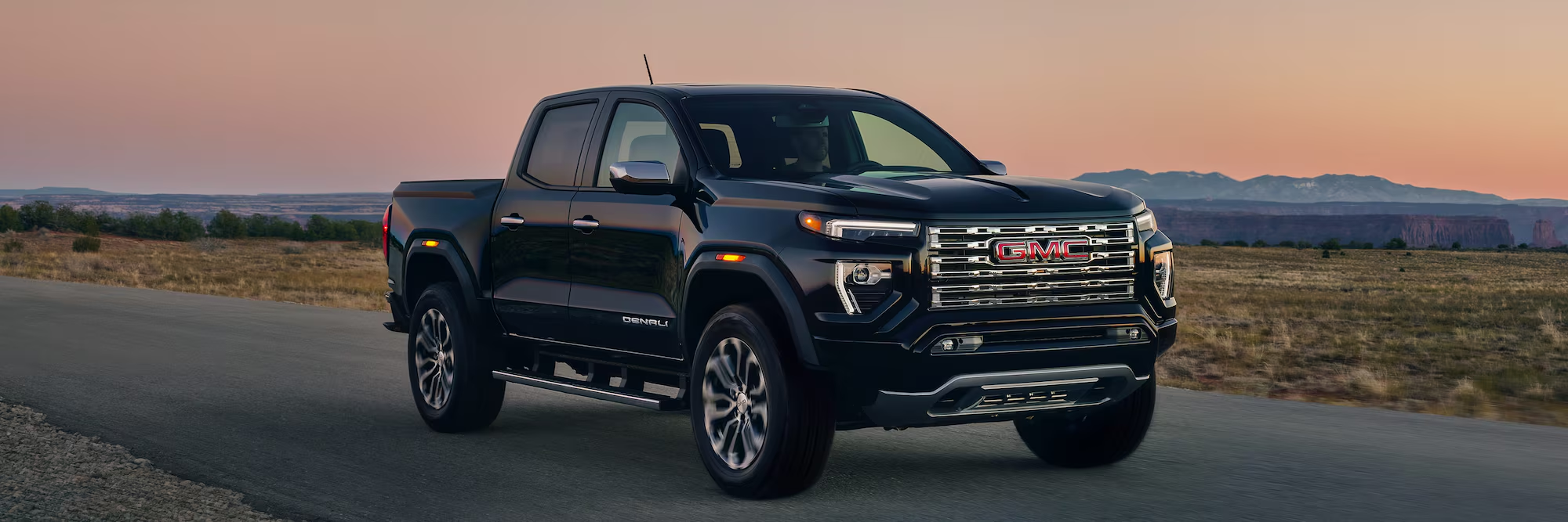 A black 2026 GMC Canyon drives along a country road at sunset