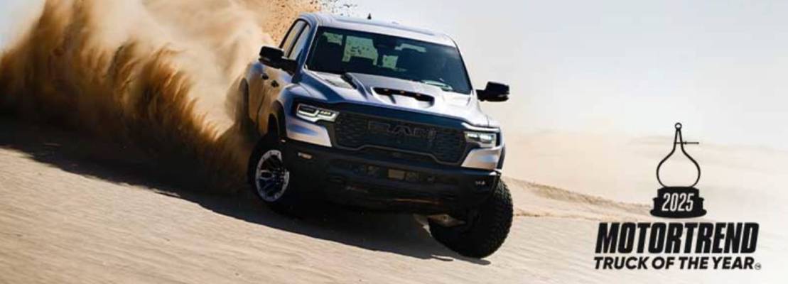 A white 2025 Ram 1500 kicks up sand as it rides over the dunes. To its right, black text declares it "2025 MotorTrend Truck of the Year".