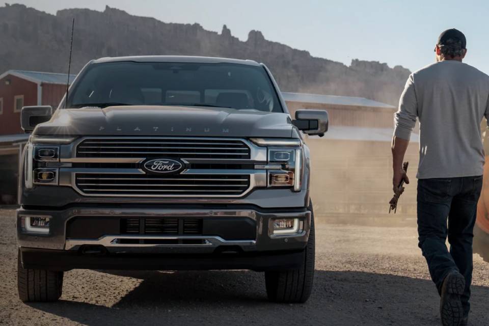 A man facing away from the camera walks past a tractor towards a waiting silver 2025 Ford F-150. Dust blows in the wind, obscuring the ranch building ahead of him.