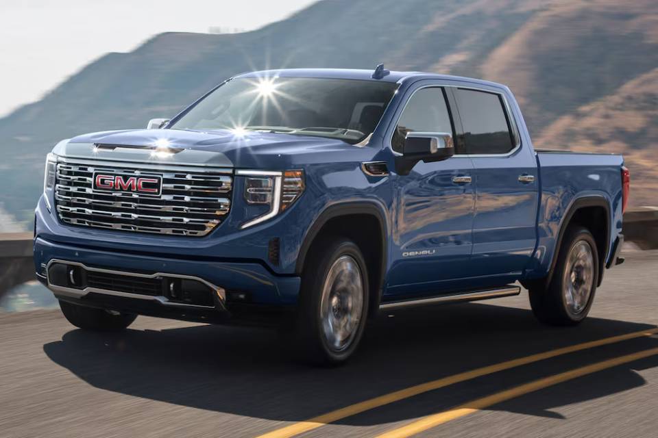 A dark blue 2025 GMC Sierra 1500 truck speeds across a bridge in front of a mountain backdrop with a river underneath.