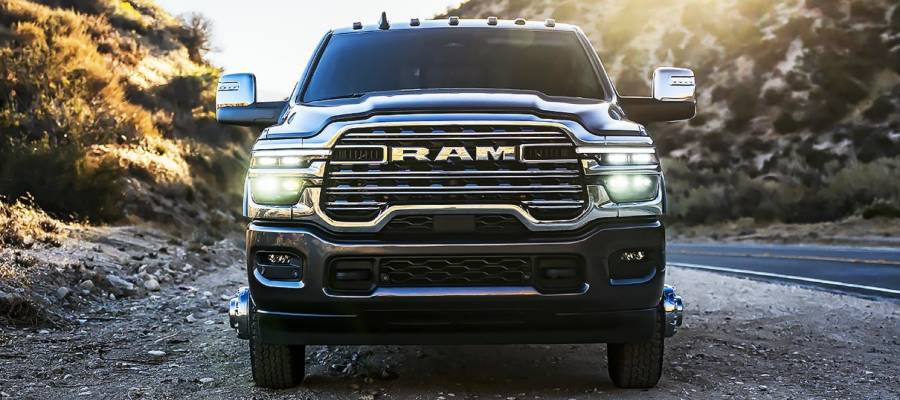 The front-on close-up of a 2025 Ram HD truck that is parked on the gravel side of a highway between trees and cliffs on either side.