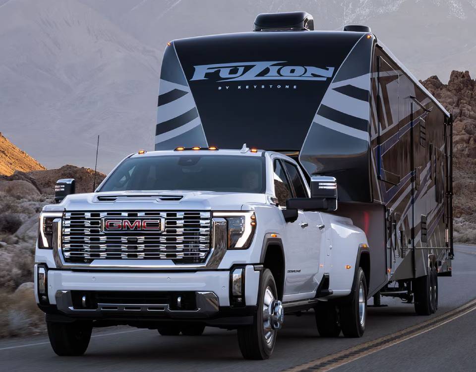 A white 2025 GMC Sierra HD truck pulls a black and silver striped 'Fuzion' trailer on a mountain road.