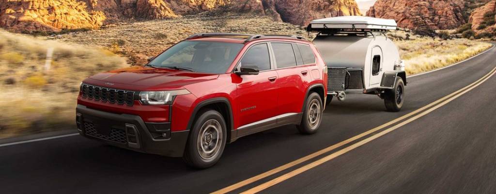 a red 2026 Jeep Cherokee pulls a silver trailer on a road past rocky cliffs