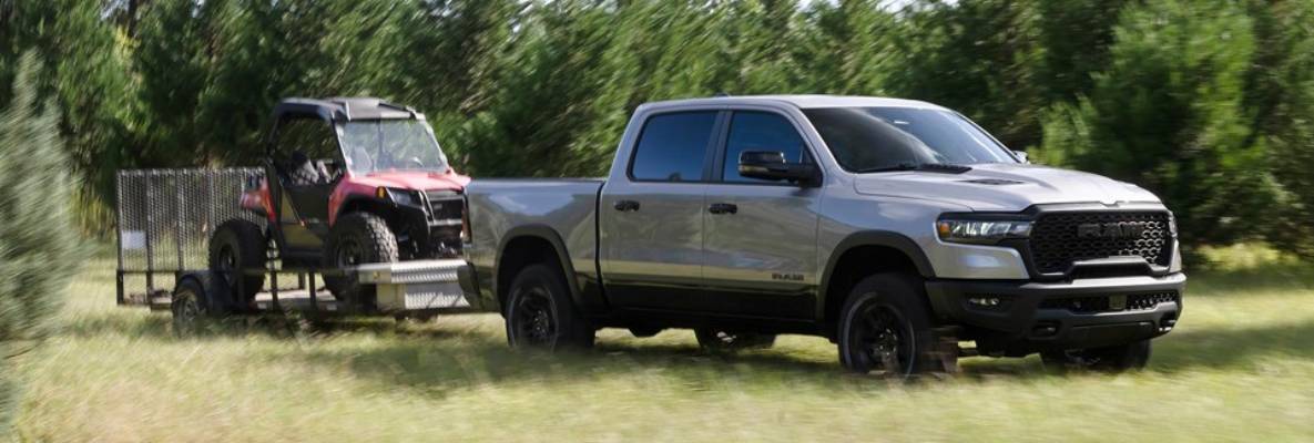 A silver 2025 ram 1500 pulls a red vehicle on a trailer across a field bordered by green trees