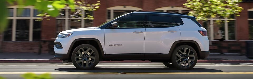 A side view of a white 2026 Jeep Compass as it drives past a brown brick building