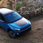 A blue 2026 Jeep Compass with available black hood insert navigates a tightly coiled dirt road uphill