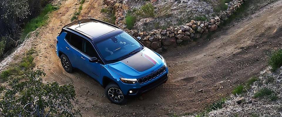A blue 2026 Jeep Compass with available black hood insert navigates a tightly coiled dirt road uphill