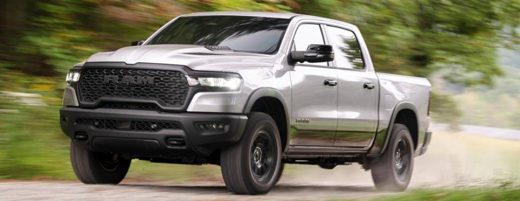 A silver 2026 Ram 1500 speeds over dirt road, kicking up clouds behind it. The trees in the background are a blur.