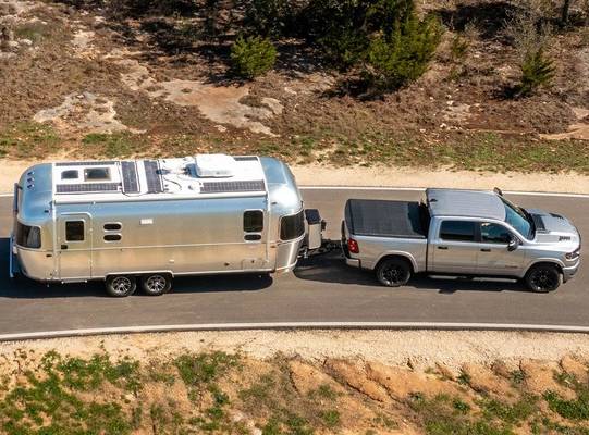 A silver 2026 Ram 1500 tows a silver trailer along a one-lane road between two banks of rough terrain