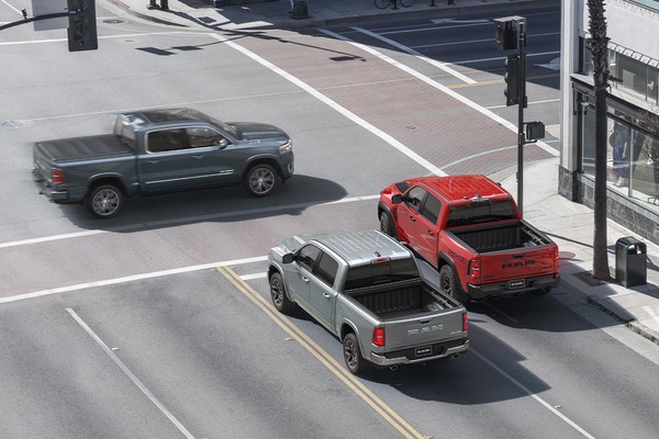 A red and a silver 2026 Ram 1500 wait at an intersection. A grey 2026 Ram 1500 passes in front of them from the left of the screen.