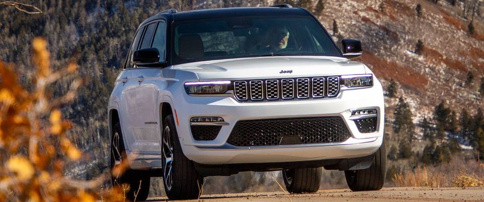 A white 2025 Jeep Grand Cherokee with a black roof drives towards the camera past a hill decked out in fall colours