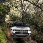 A white 2026 Jeep Compass with a black roof and hood spoiler drives towards the camera through a forest