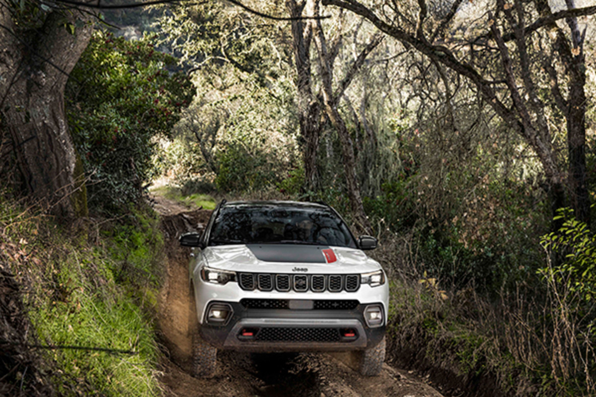 A white 2026 Jeep Compass with a black roof and hood spoiler drives towards the camera through a forest