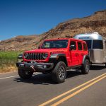 A red 2026 Jeep Wrangler tows a silver camper along a road beside green grass and brown cliff
