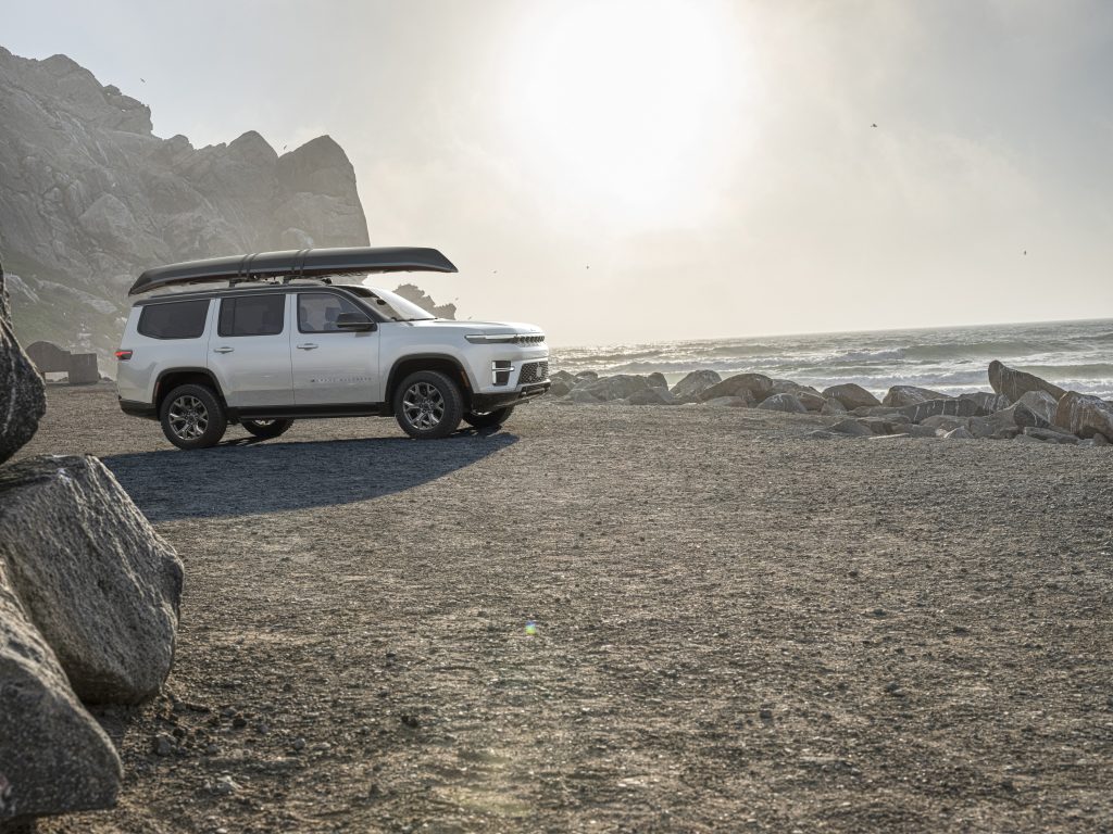 2026 Jeep Grand Wagoneer white full‑size SUV parked on a rocky shoreline with a canoe mounted on the roof, lifestyle image used by Leavens Chrysler Dodge Jeep RAM London Ontario.